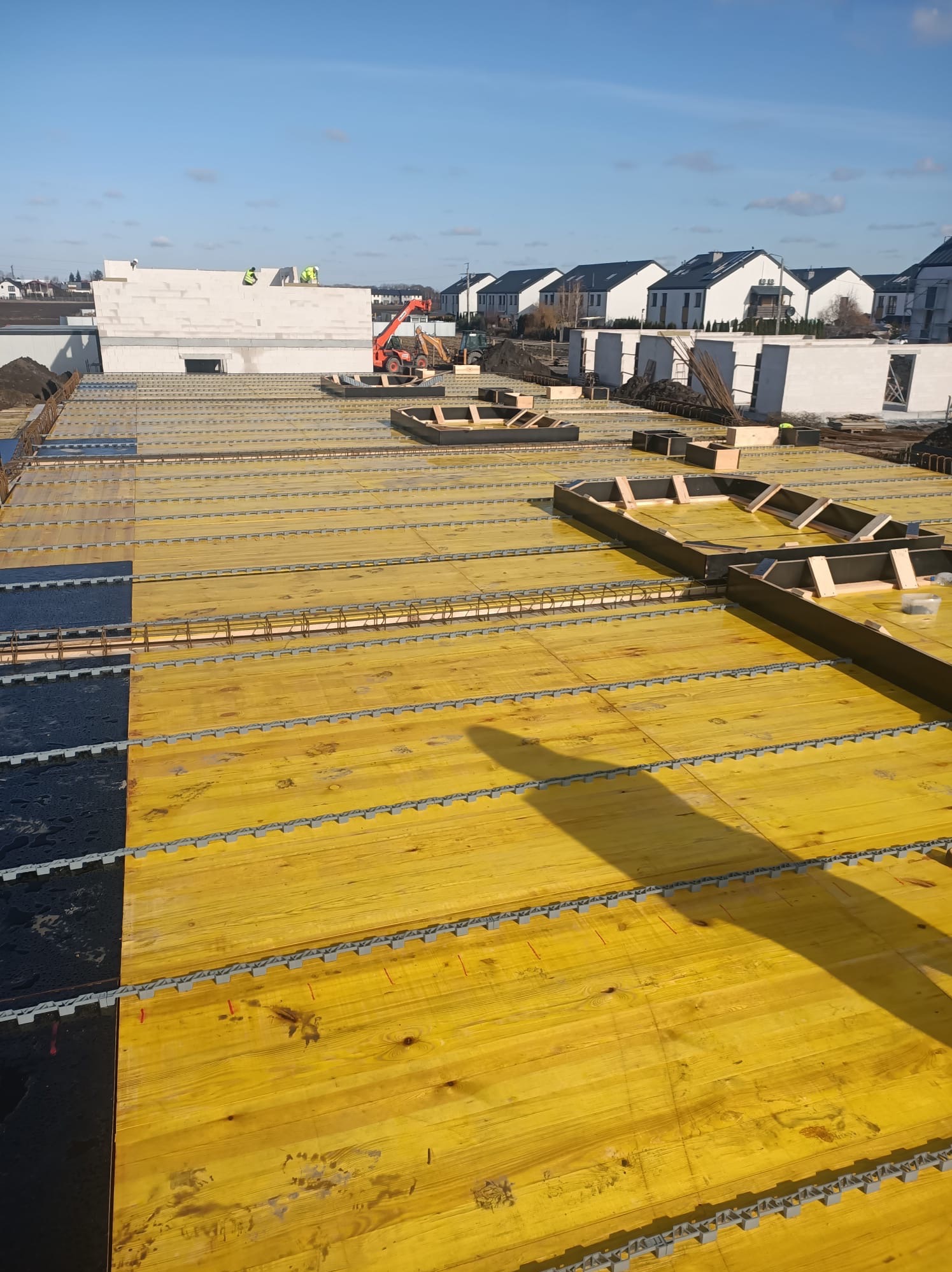 Żółte szalunki stropowe zbrojone stalą na budowie domu, w tle budynki mieszkalne i pracujący dźwig. Widok z góry, w słoneczny dzień.