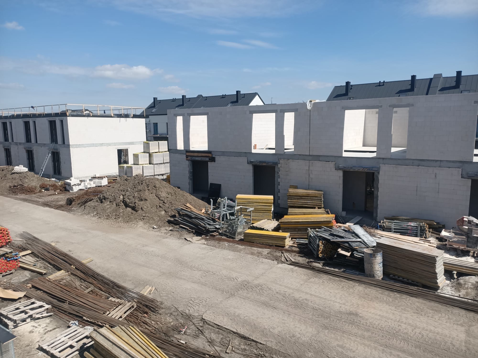 Budowa domów z bloczków betonu komórkowego w trakcie realizacji. Widoczne ściany, otwory okienne i drzwiowe, stosy materiałów budowlanych i zbrojenia.