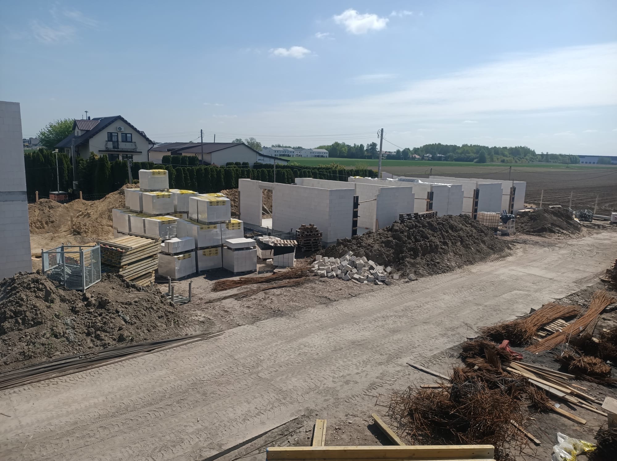 Wznoszenie ścian domu z bloczków betonu komórkowego, widoczne palety z materiałem i zbrojenie. Budowa w otoczeniu pól i zabudowy jednorodzinnej.