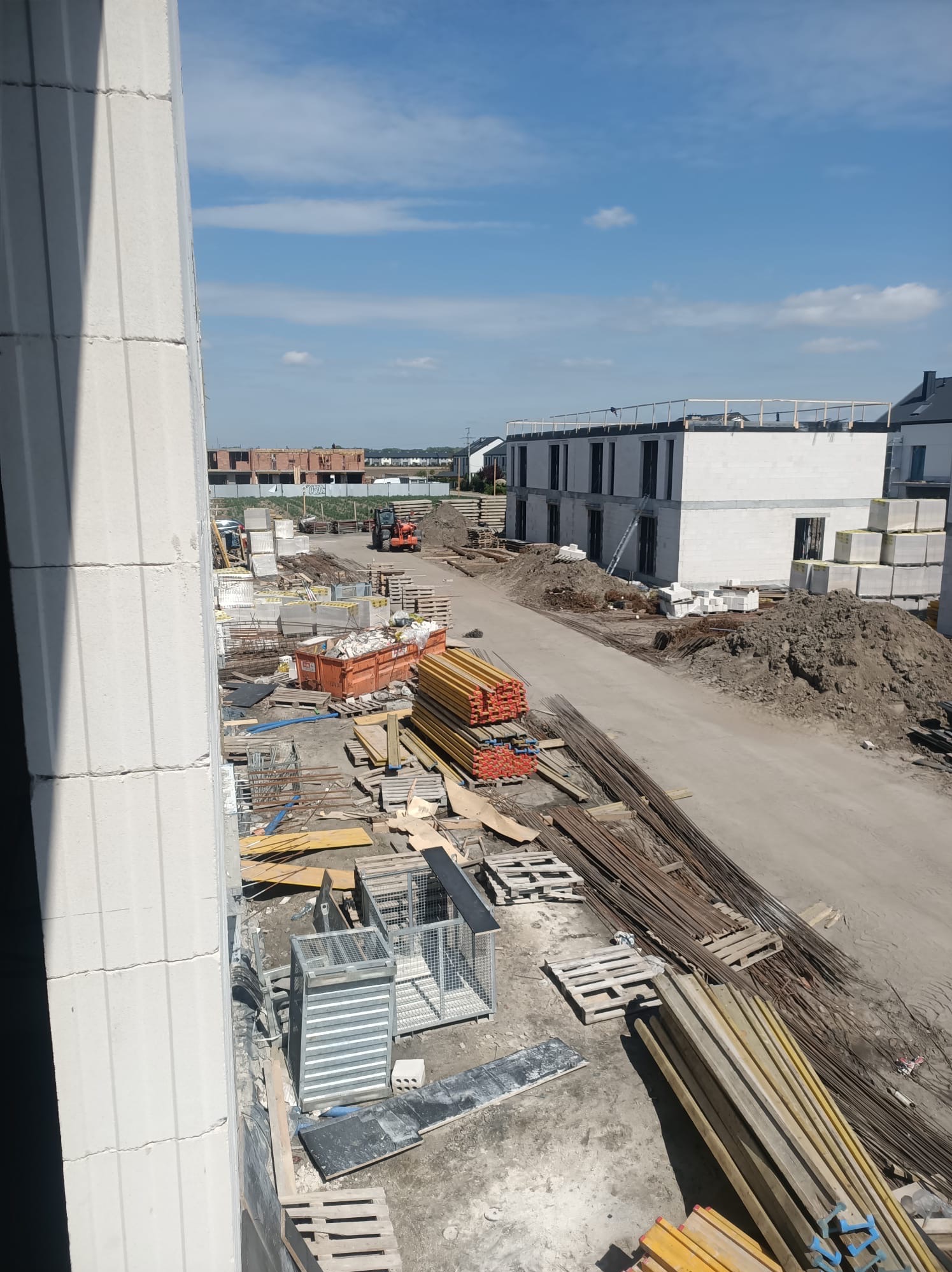 Budowa domu w trakcie wznoszenia ścian z bloczków, widoczne materiały budowlane, teren budowy z lekkim sprzętem. Ujęcie z góry, w tle budynki.