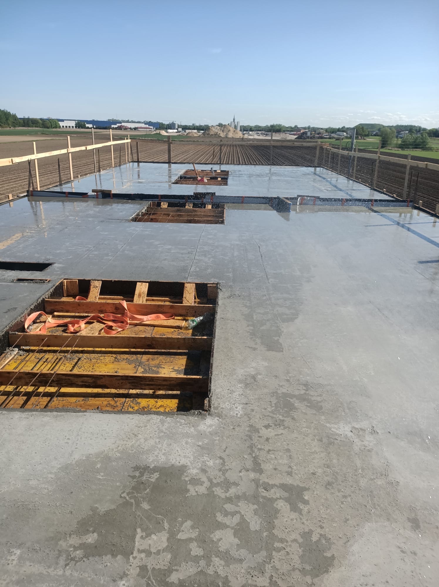 Zalany fundament z widocznym zbrojeniem i drewnianym szalunkiem. W tle pole uprawne i zabudowania.