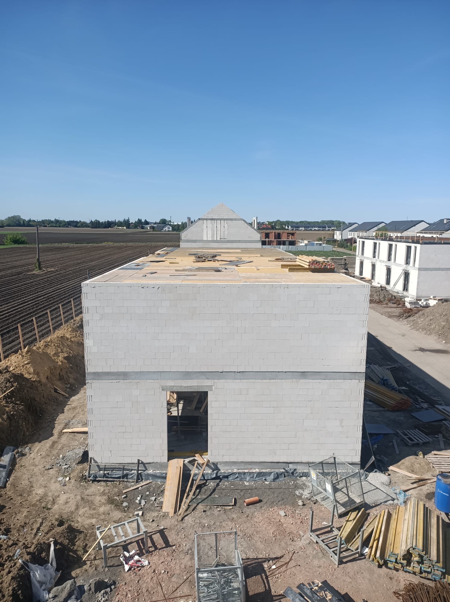 Surowy stan domu z szarej cegły silikatowej, widok z góry na plac budowy, w tle pola i zabudowa jednorodzinna. Widoczne drewniane elementy konstrukcji dachu.