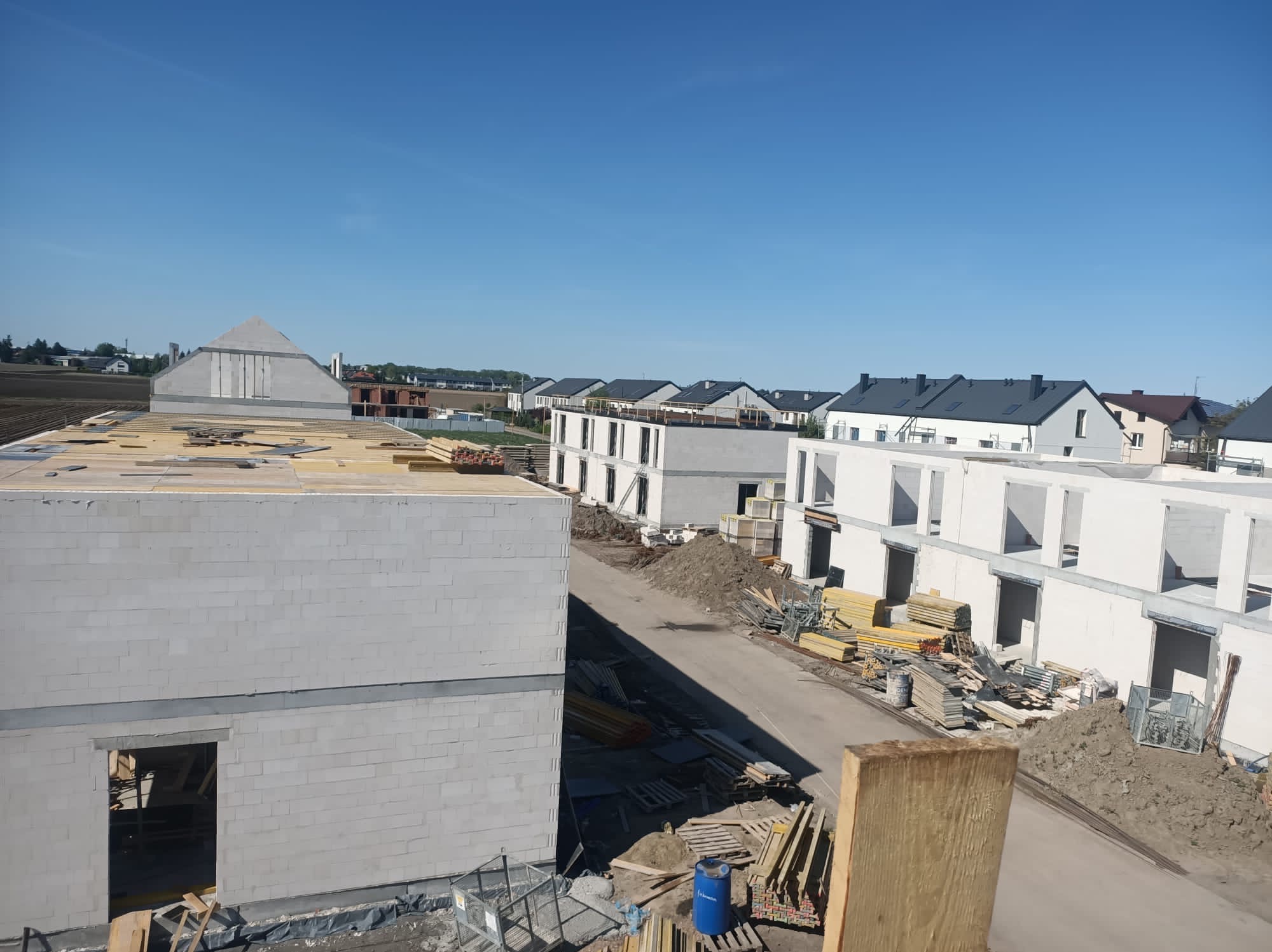 Wysoki kąt na budowę szeregowców z białego bloczka, widoczne ściany i stropy w stanie surowym otwarte, materiały budowlane i droga dojazdowa. Błękitne niebo.