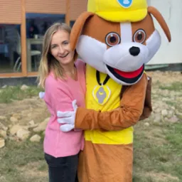 Uśmiechnięta blondynka w różowym swetrze obejmuje pluszową maskotkę psa w żółtym kasku i ubraniu, na zewnątrz, w tle budynek z brązowymi żaluzjami.