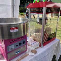 FUNNY KIDS Agnieszka Góra - Maszyna do waty cukrowej w kolorze różowym obok maszyny do popcornu w czerwonej obudowie, ustawione na białym stole pod namiotem, z widocznymi akcesoriami do pakowania.