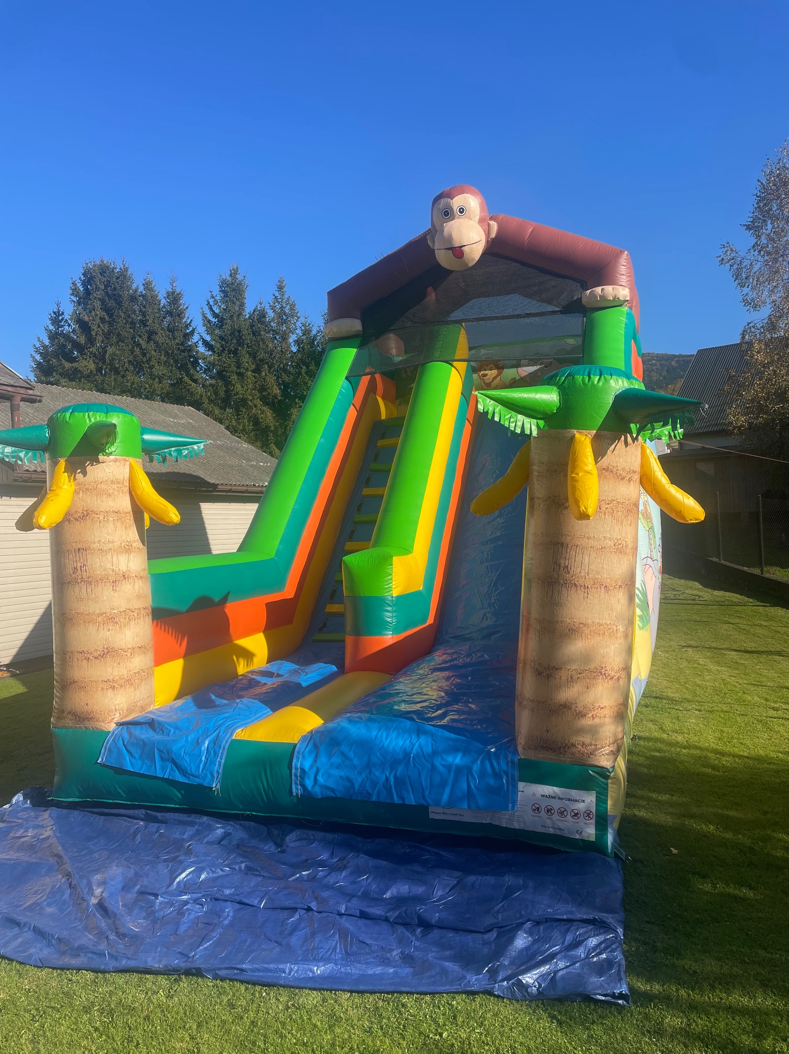 Kolorowy dmuchany zamek ze zjeżdżalnią w motywie dżungli z małpą na szczycie i palmami po bokach, rozłożony na trawniku na niebieskiej plandece.