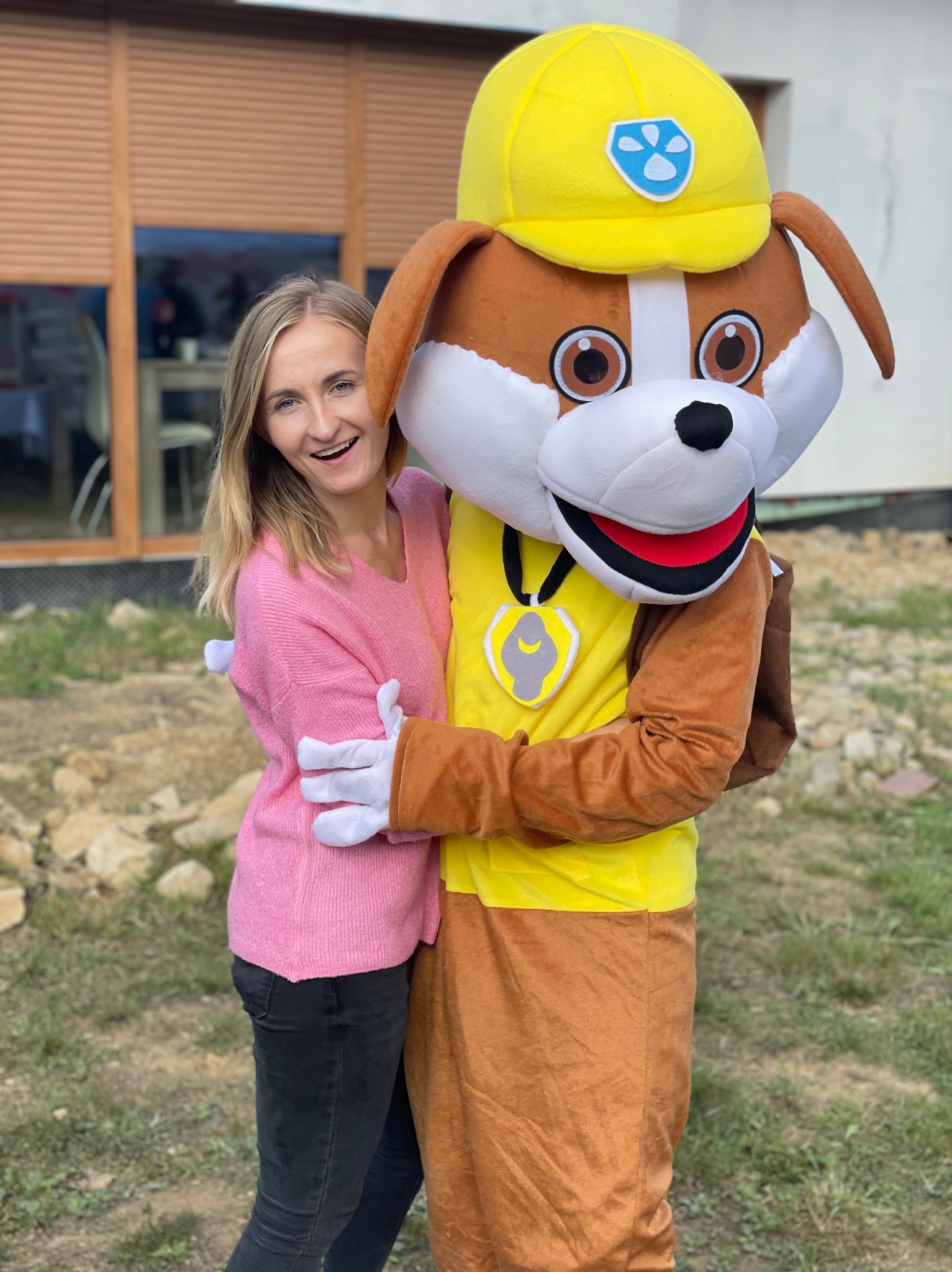 Uśmiechnięta blondynka w różowym swetrze obejmuje pluszową maskotkę psa w żółtym kasku i ubraniu, na zewnątrz, w tle budynek z brązowymi żaluzjami.