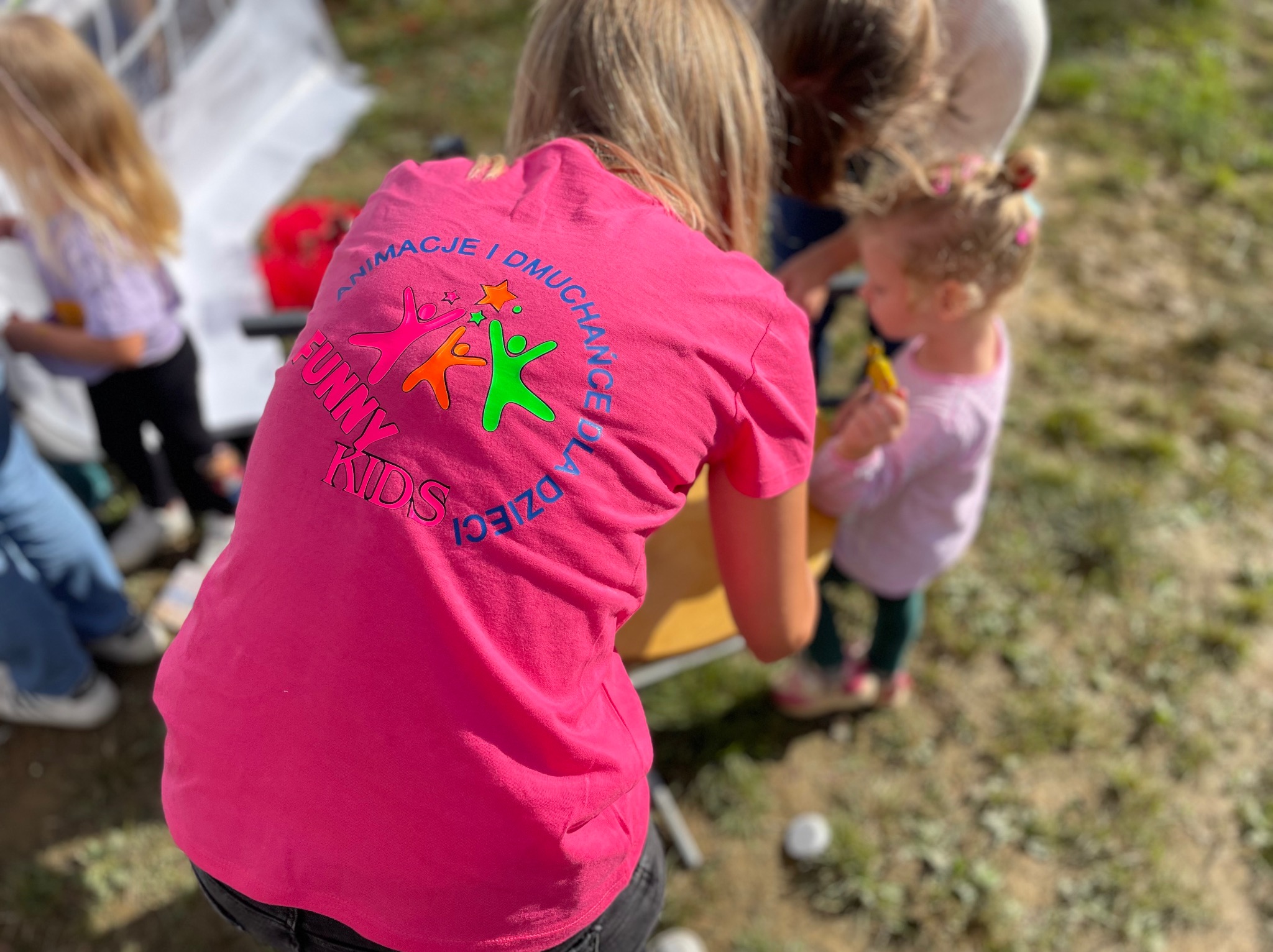 Kobieta w różowej koszulce z napisem 'Animacje i Dmuchańce dla Dzieci Funny Kids' w otoczeniu dzieci, prawdopodobnie podczas organizacji imprezy plenerowej.