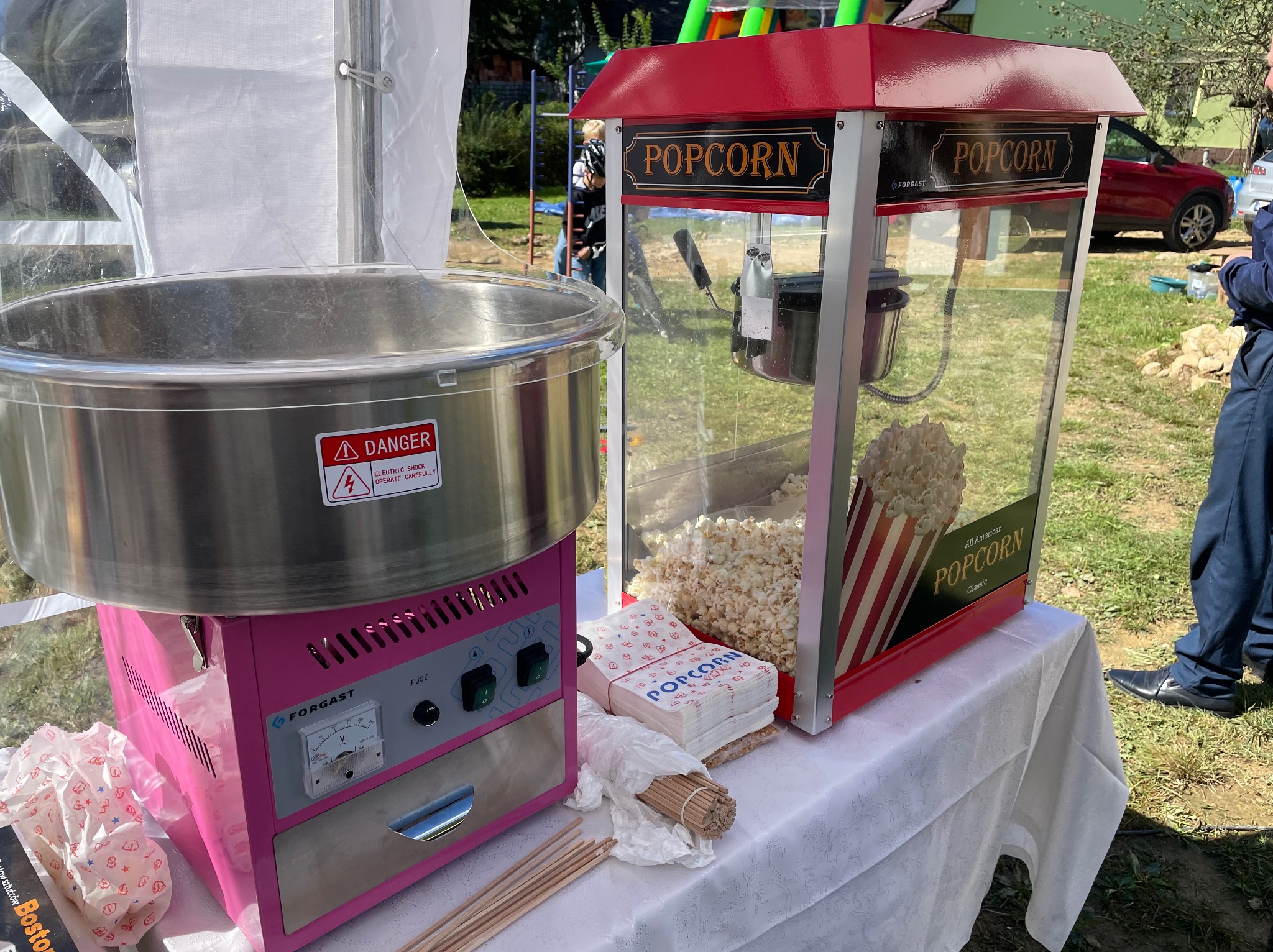 Maszyna do waty cukrowej w kolorze różowym obok maszyny do popcornu w czerwonej obudowie, ustawione na białym stole pod namiotem, z widocznymi akcesoriami do pakowania.