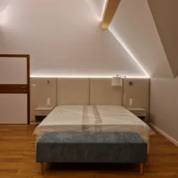 Beżowe łóżko z zagłówkiem w pokoju na poddaszu, oświetlenie LED, drewniana belka. Nowoczesny wystrój sypialni z drewnianą podłogą i tapicerowanym siedziskiem.