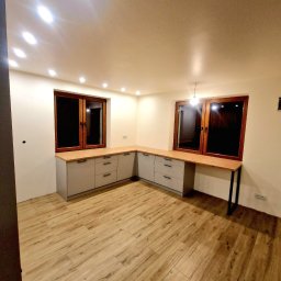 F.H.U KRZYSZTOF KLIMCZAK - Szare szafki kuchenne z drewnianym blatem pod oknami, nowoczesne oświetlenie sufitowe, panele podłogowe imitujące drewno, widok z perspektywy narożnika pomieszczenia.