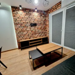 Salon z ceglaną ścianą, drewnianą szafką RTV i stolikami kawowymi. Nowoczesne oświetlenie i żaluzje okienne. Ciepłe odcienie drewna i cegły.