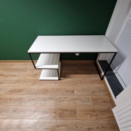 F.H.U KRZYSZTOF KLIMCZAK - Minimalistyczne biurko z białym blatem i czarnymi nogami, z półkami po lewej stronie, na tle zielonej ściany i drewnianej podłogi. Nowoczesny design.