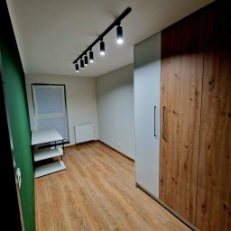 F.H.U KRZYSZTOF KLIMCZAK - Nowoczesna szafa z drewnianymi frontami i szarym bokiem, czarne uchwyty, oświetlenie punktowe na suficie, parkiet i minimalistyczny regał w pokoju.