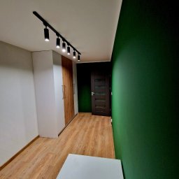 F.H.U KRZYSZTOF KLIMCZAK - Wnętrze z szafą na wymiar o białych frontach i drewnianych akcentach, oświetloną szynoprzewodem, na tle zielonej ściany i drewnianej podłogi.