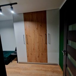 F.H.U KRZYSZTOF KLIMCZAK - Nowoczesna szafa z frontami łączącymi biel i dekor drewna, z długimi, czarnymi uchwytami, na tle drewnianej podłogi i fragmentu zielonej ściany.