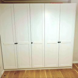 F.H.U KRZYSZTOF KLIMCZAK - Biała szafa z sześcioma drzwiami z czarnymi uchwytami, na drewnianej podłodze. Uwagę przyciąga minimalistyczny design i dopasowanie do wnętrza.