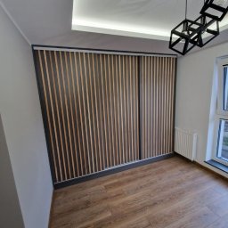 F.H.U KRZYSZTOF KLIMCZAK - Nowoczesna szafa wnękowa z przesuwnymi drzwiami lamelowymi w jasnym wnętrzu. Widoczne drewniane panele i minimalistyczna lampa.