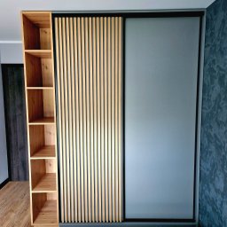 F.H.U KRZYSZTOF KLIMCZAK - Nowoczesna szafa wnękowa z przesuwnymi drzwiami: matowe szkło i drewniane lamele. Obok regał z jasnego drewna. Minimalistyczny design, szara ściana w tle.