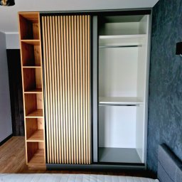 F.H.U KRZYSZTOF KLIMCZAK - Szafa wnękowa z przesuwnymi drzwiami z drewnianymi lamelami, z półkami z boku, na tle szarej ściany w sypialni, widok z dołu.