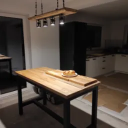 Drewniany stół barowy z czarnymi nogami i oświetleniem wiszącym w kuchni. Na blacie leży chałka na talerzu. Widoczne białe szafki kuchenne w tle.