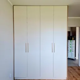 Czterodrzwiowa, biała szafa z drewnianymi obramowaniami i podłużnymi, metalowymi uchwytami, ustawiona na drewnianej podłodze w jasnym wnętrzu.