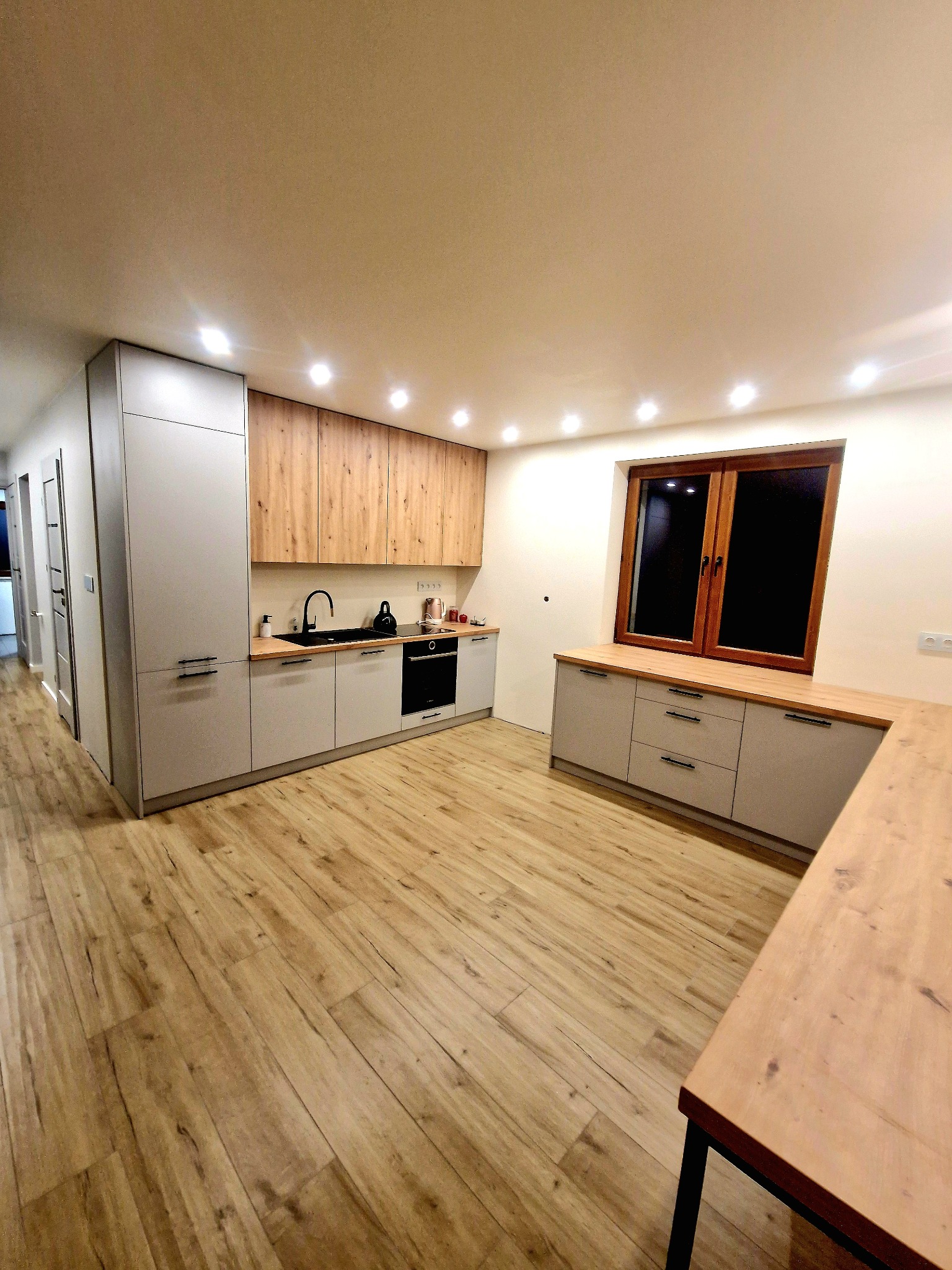 Nowoczesna kuchnia z szarymi szafkami dolnymi i drewnianymi górnymi. Blat z drewna pod oknem. Jasna podłoga imitująca drewno. Oświetlenie punktowe sufitowe.