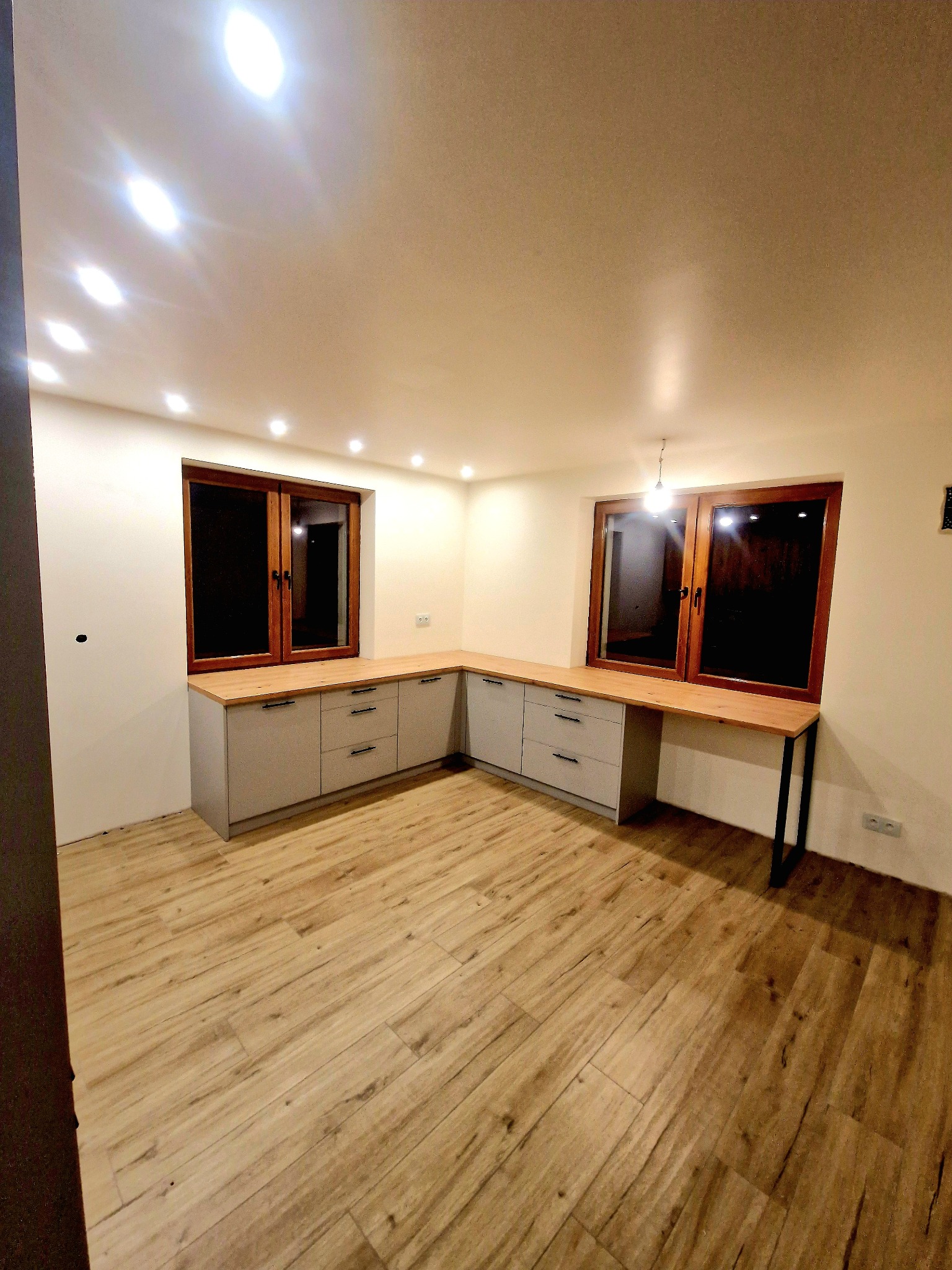 Szare szafki kuchenne z drewnianym blatem pod oknami, nowoczesne oświetlenie sufitowe, panele podłogowe imitujące drewno, widok z perspektywy narożnika pomieszczenia.