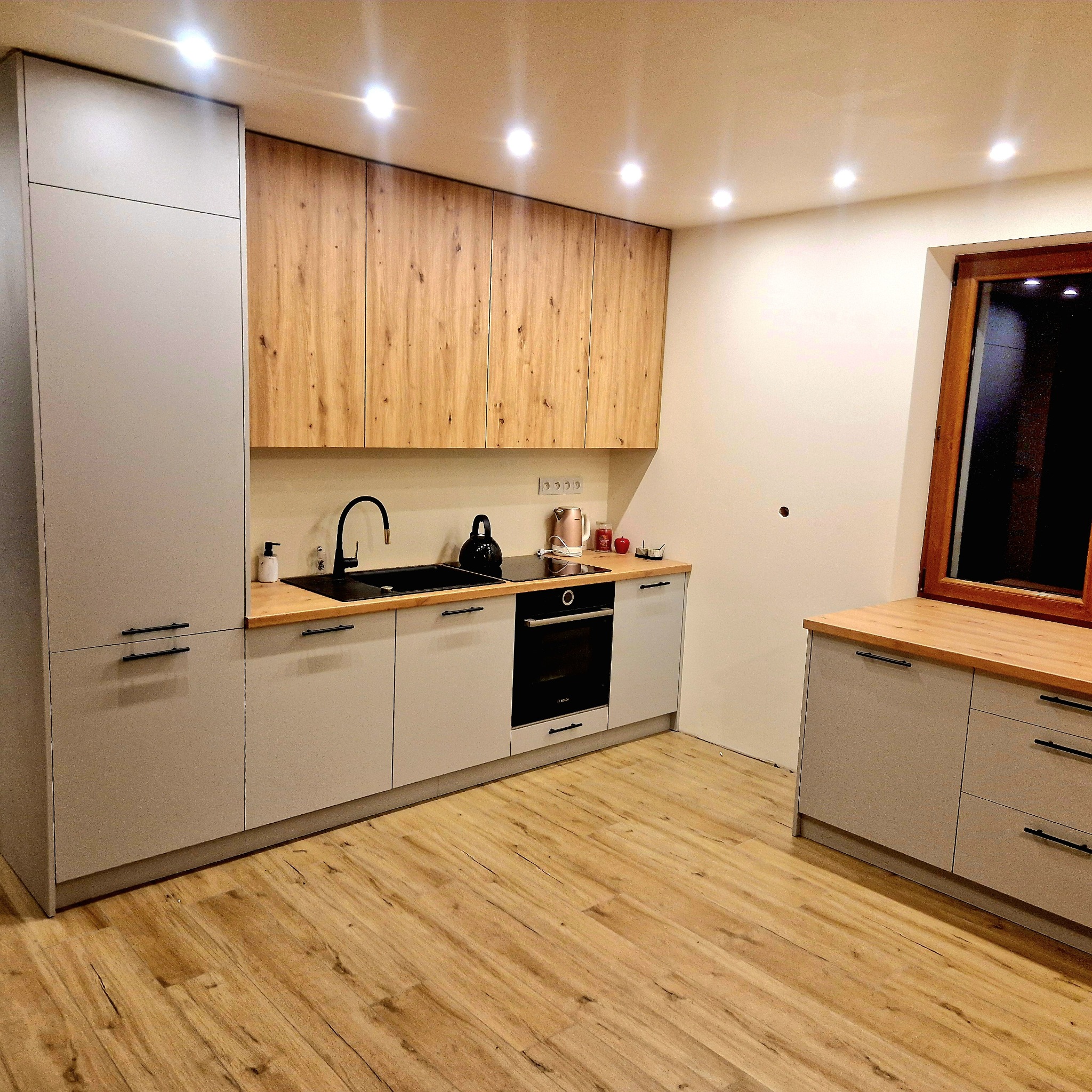 Nowoczesna kuchnia w Krościenku nad Dunajcem. Szare szafki dolne, drewnopodobne górne. Czarny zlew i bateria. Jasna podłoga imitująca drewno.