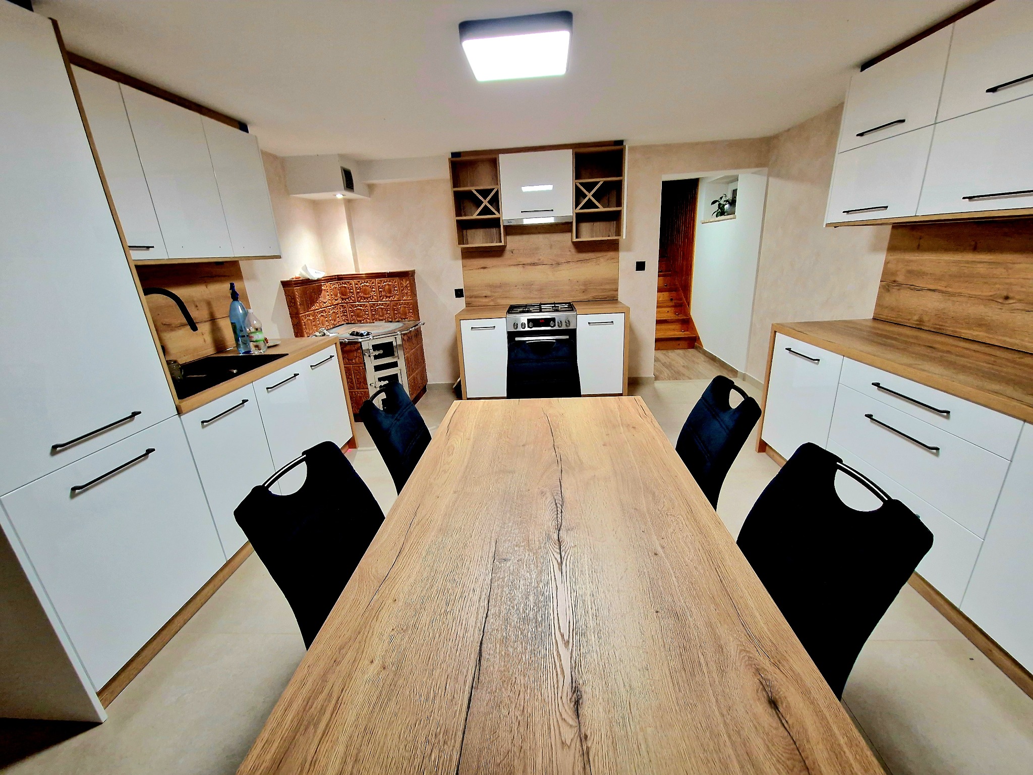 Nowoczesna kuchnia z białymi szafkami i drewnianym stołem. Widoczny piec kaflowy i drewniane wykończenia. Jasne wnętrze z oświetleniem LED.