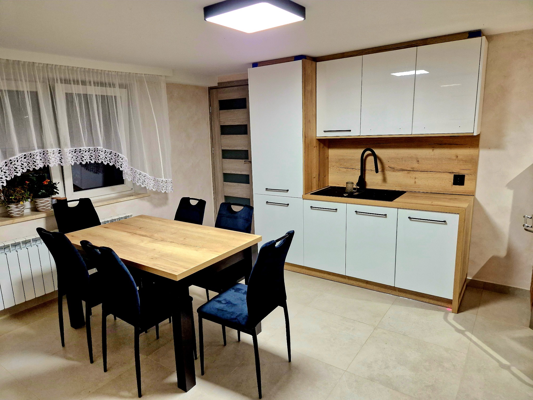 Nowoczesna kuchnia z białymi frontami i drewnianym blatem, uzupełniona o stół z krzesłami w odcieniach granatu, całość oświetlona ciepłym światłem.