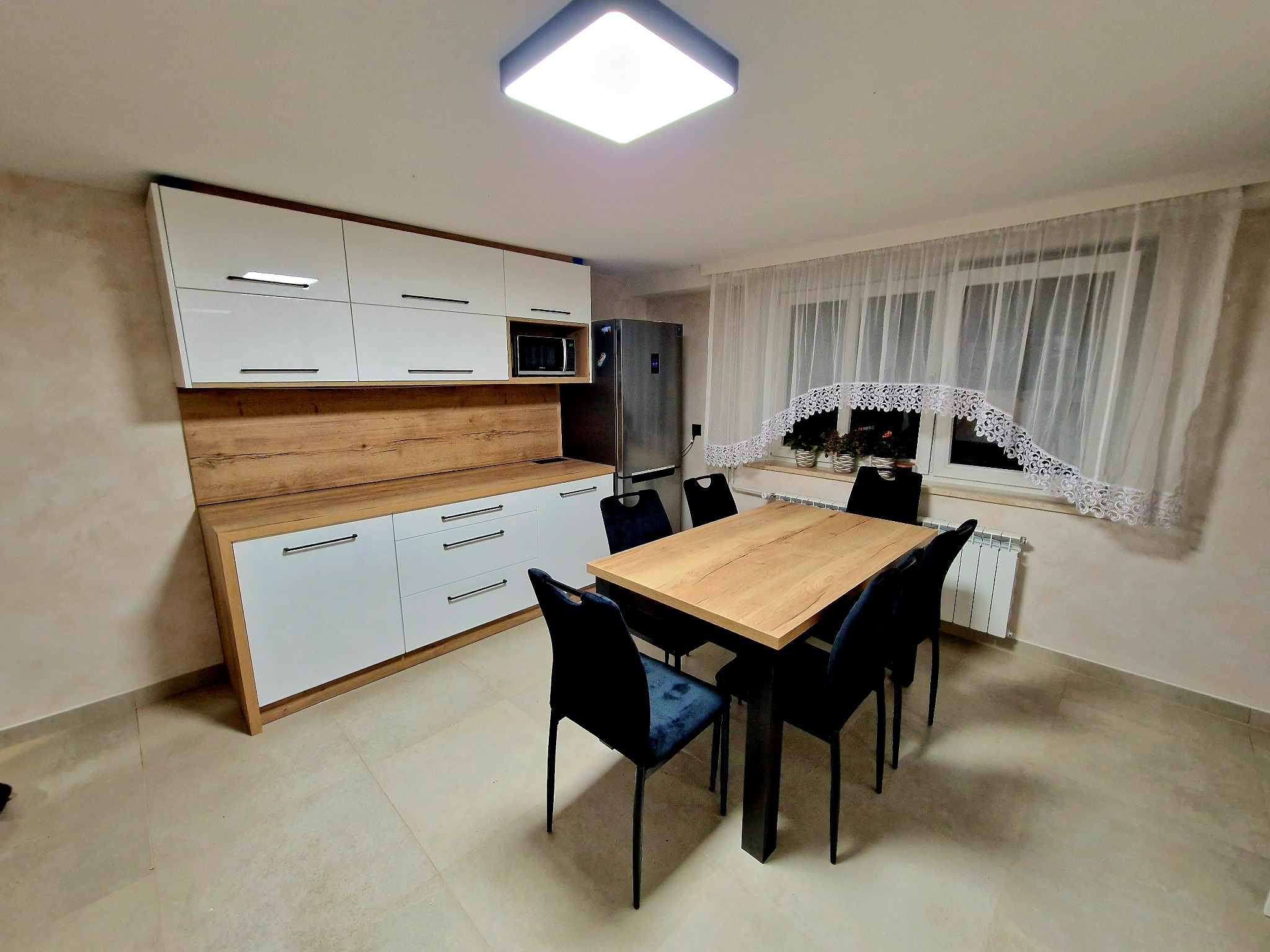 Jasna kuchnia z białymi szafkami i drewnianym blatem, stół z ciemnymi krzesłami. Nowoczesny design wnętrza, widoczne okno z firanką i grzejnik.