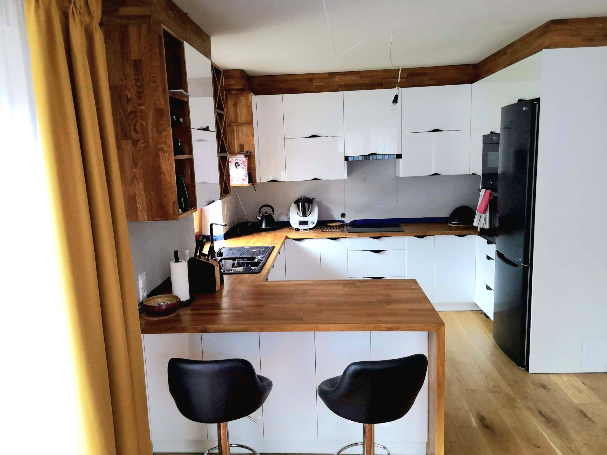 Nowoczesna kuchnia z białymi szafkami i drewnianym blatem, uzupełniona o hokerowe krzesła i złote zasłony, tworząca spójną całość z drewnianym wykończeniem sufitu.