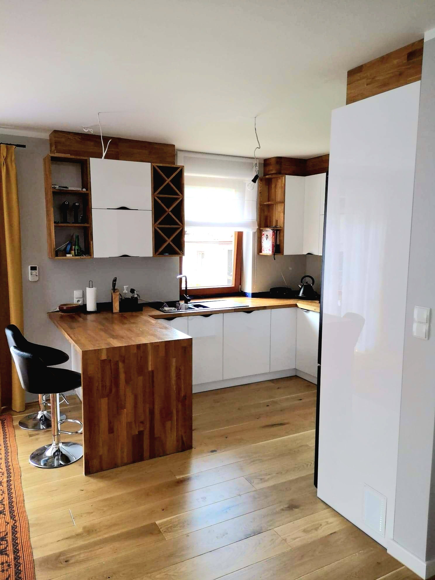 Jasna kuchnia z drewnianym blatem i barkiem. Białe fronty szafek, drewniane półki i regał na wino. Parkiet na podłodze, dywanik z etnicznym wzorem.