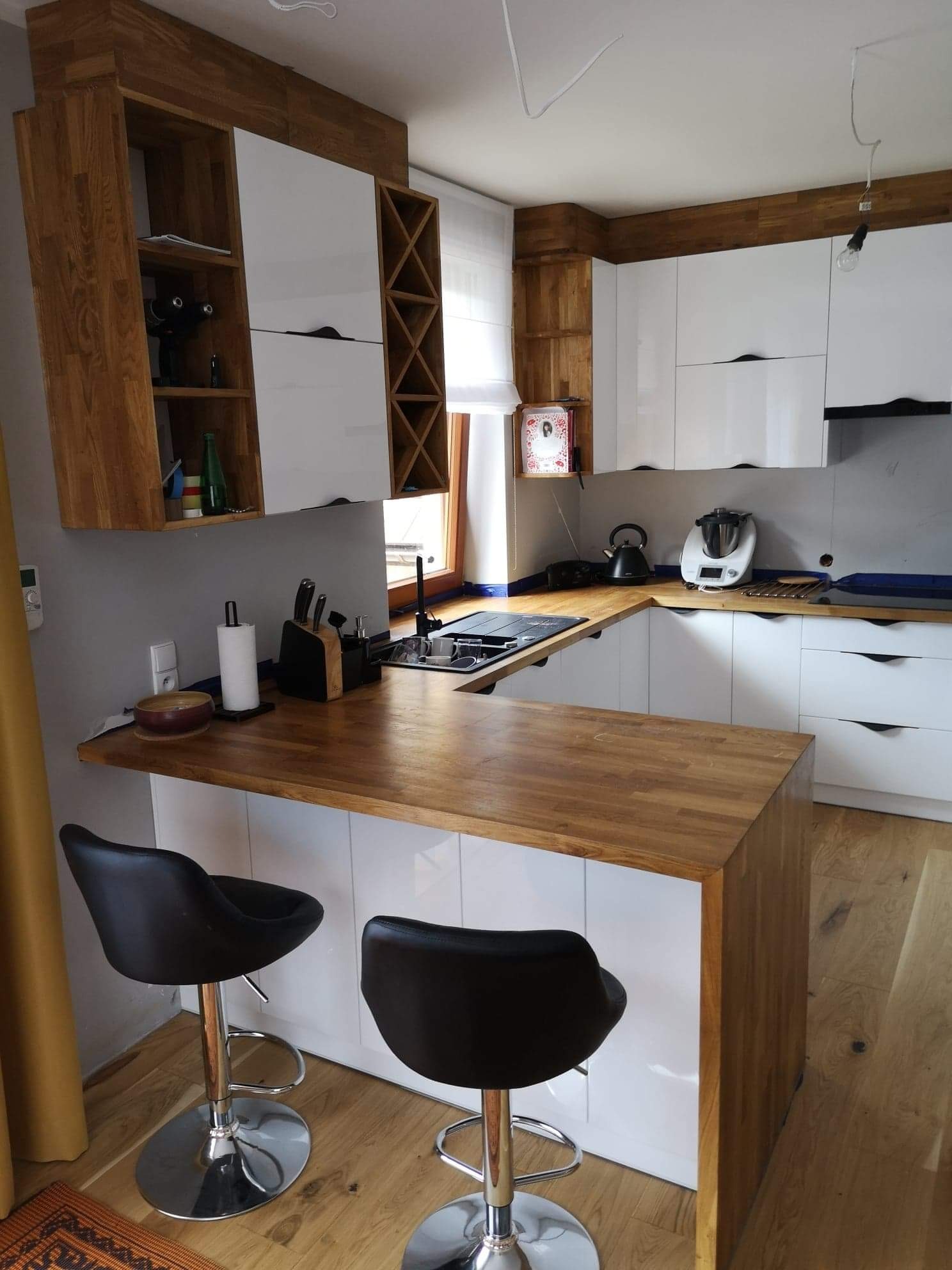 Nowoczesna kuchnia z białymi szafkami i drewnianym blatem. Wyspa kuchenna z hokerami. Oświetlenie punktowe i naturalne światło z okna. Elementy drewniane na górnych szafkach.