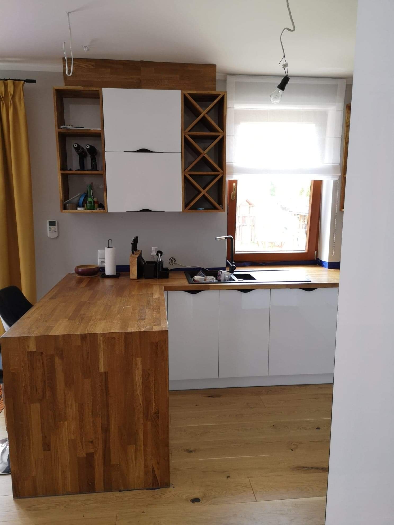 Jasna kuchnia na wymiar z drewnianym blatem i barkiem. Białe fronty szafek i drewniane akcenty. Widoczne wyposażenie i okno z roletą rzymską.