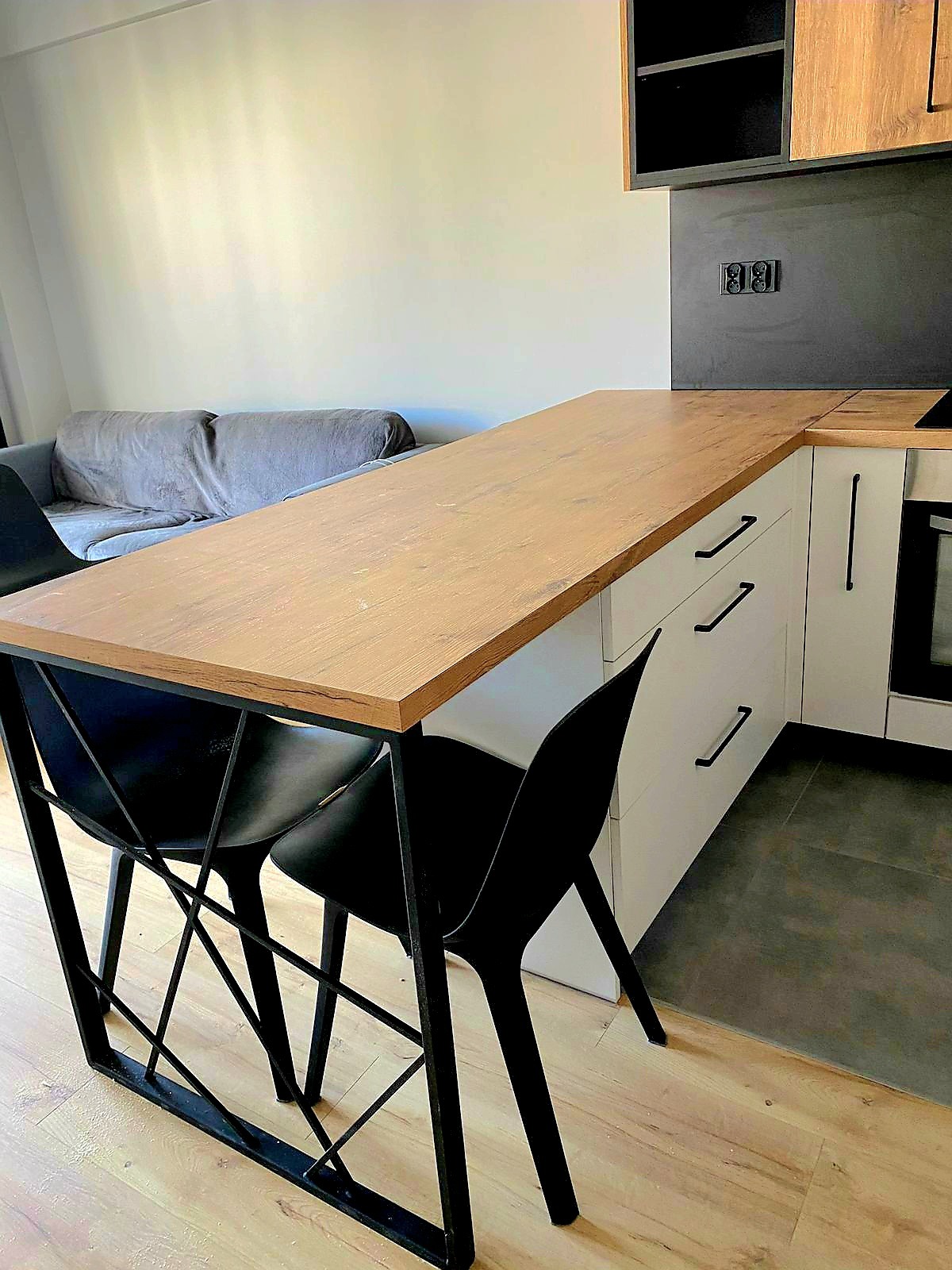 Nowoczesna kuchnia z drewnianym blatem, białymi szafkami i czarnymi uchwytami. Dwa czarne krzesła z metalową podstawą. W tle szara sofa i czarny panel nad blatem.