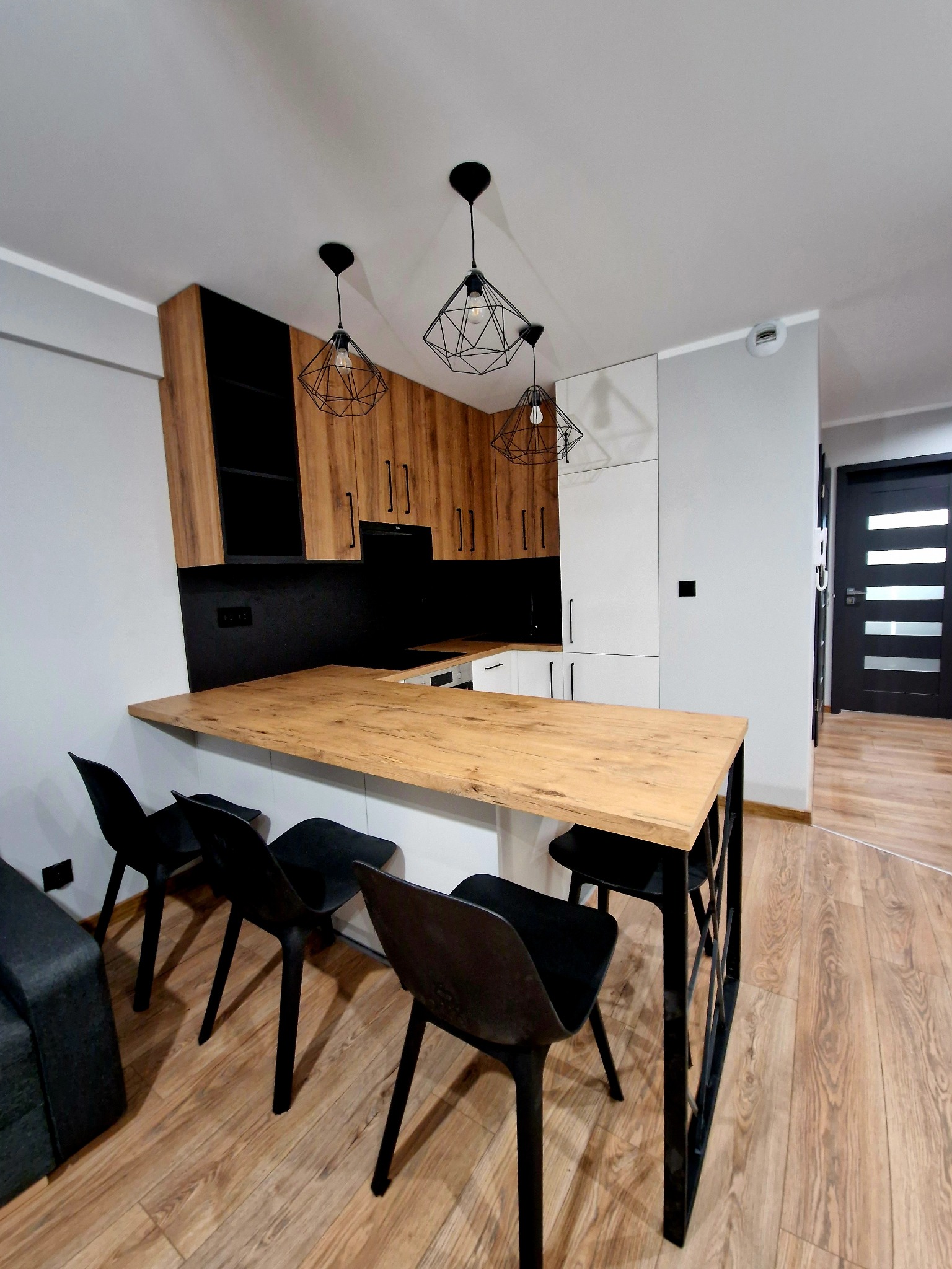 Nowoczesna kuchnia z drewnianymi szafkami, czarnym blatem i wyspą z drewnianym blatem oraz czarnymi krzesłami. Nad wyspą wiszą designerskie, geometryczne lampy.