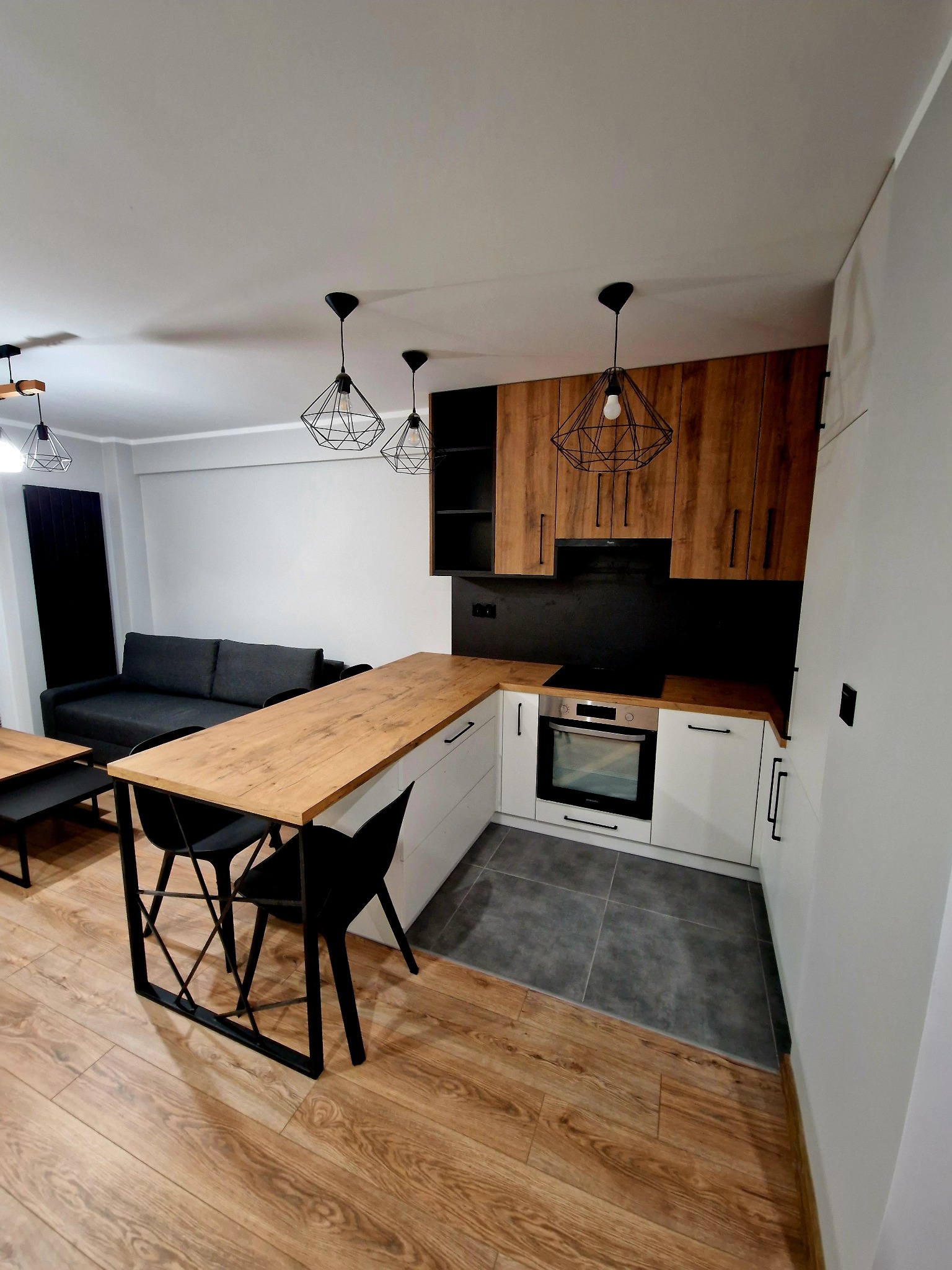 Nowoczesna kuchnia z drewnianymi szafkami górnymi i białymi dolnymi, wyspą z blatem drewnianym i czarnymi krzesłami. Lampy wiszące w geometrycznym stylu.