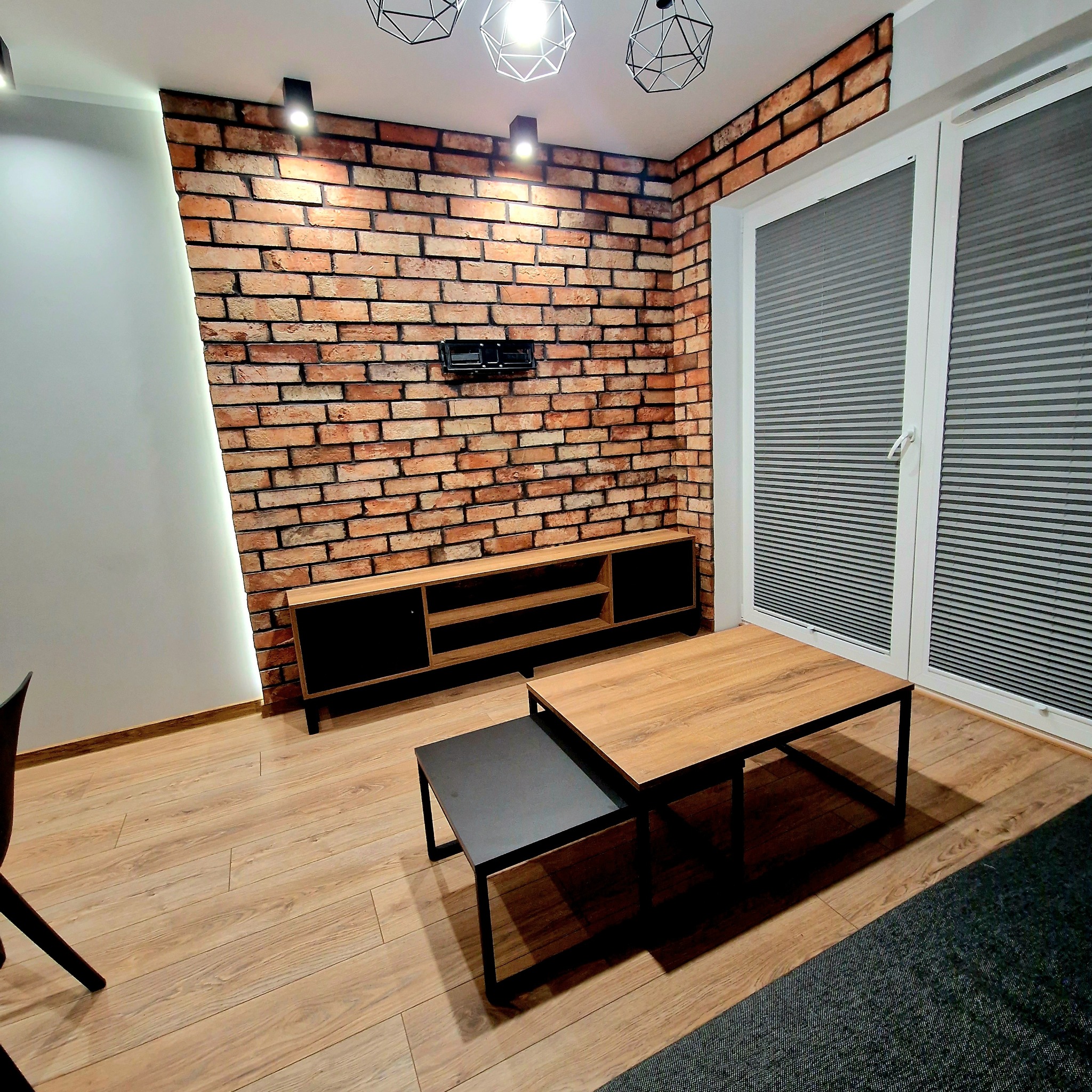 Salon z ceglaną ścianą, drewnianą szafką RTV i stolikami kawowymi. Nowoczesne oświetlenie i żaluzje okienne. Ciepłe odcienie drewna i cegły.