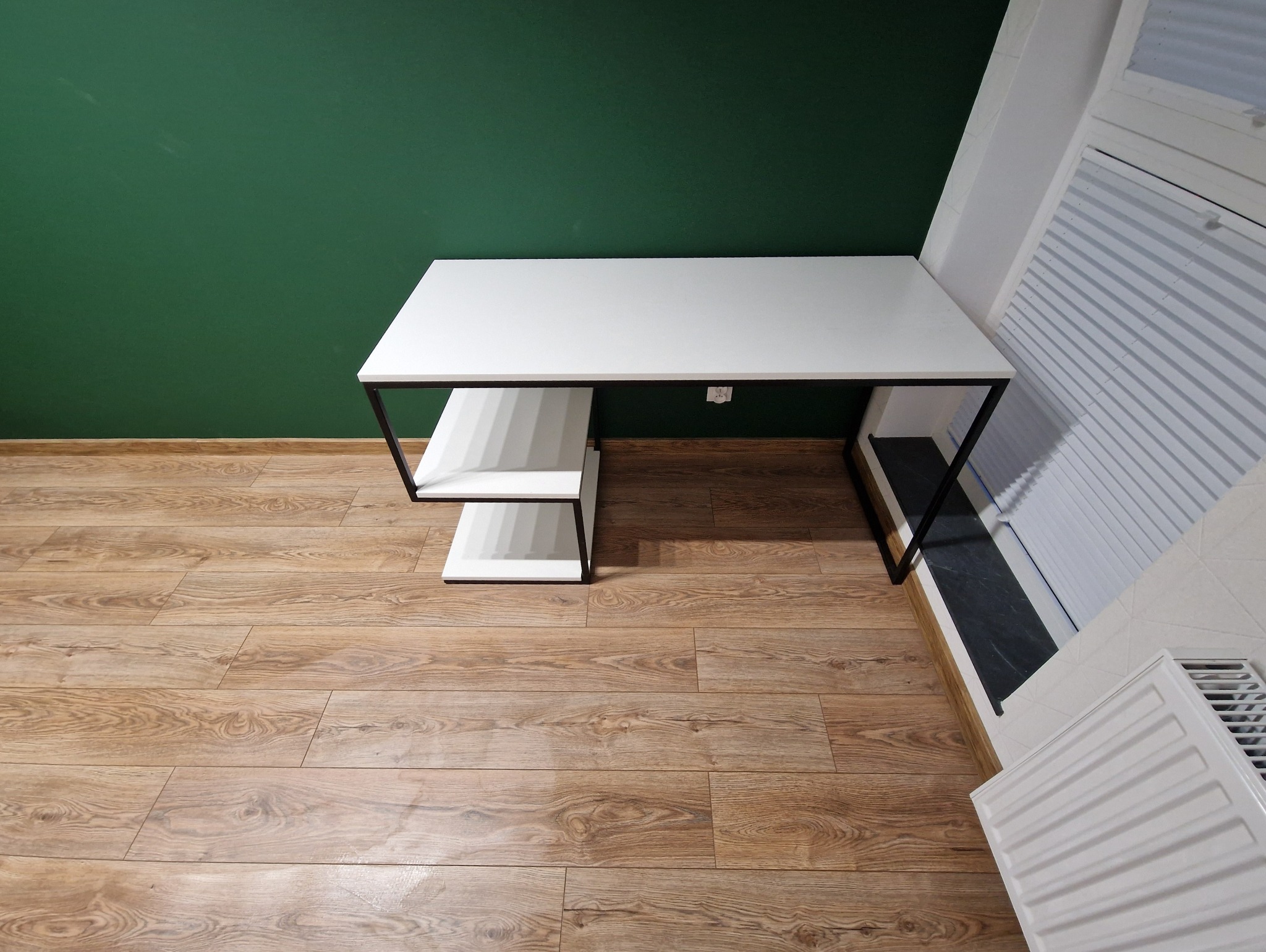 Minimalistyczne biurko z białym blatem i czarnymi nogami, z półkami po lewej stronie, na tle zielonej ściany i drewnianej podłogi. Nowoczesny design.