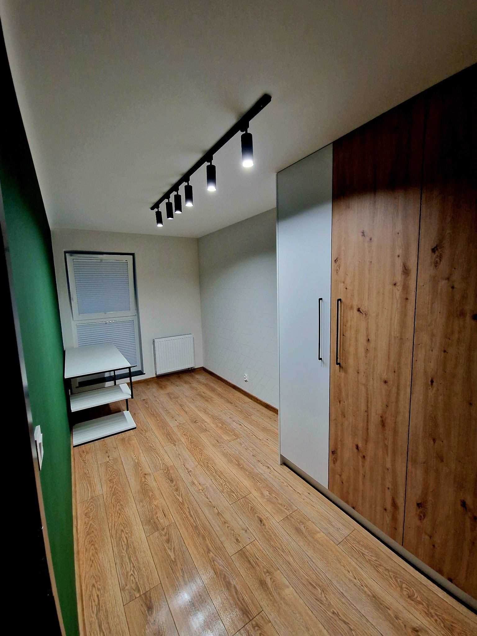 Nowoczesna szafa z drewnianymi frontami i szarym bokiem, czarne uchwyty, oświetlenie punktowe na suficie, parkiet i minimalistyczny regał w pokoju.