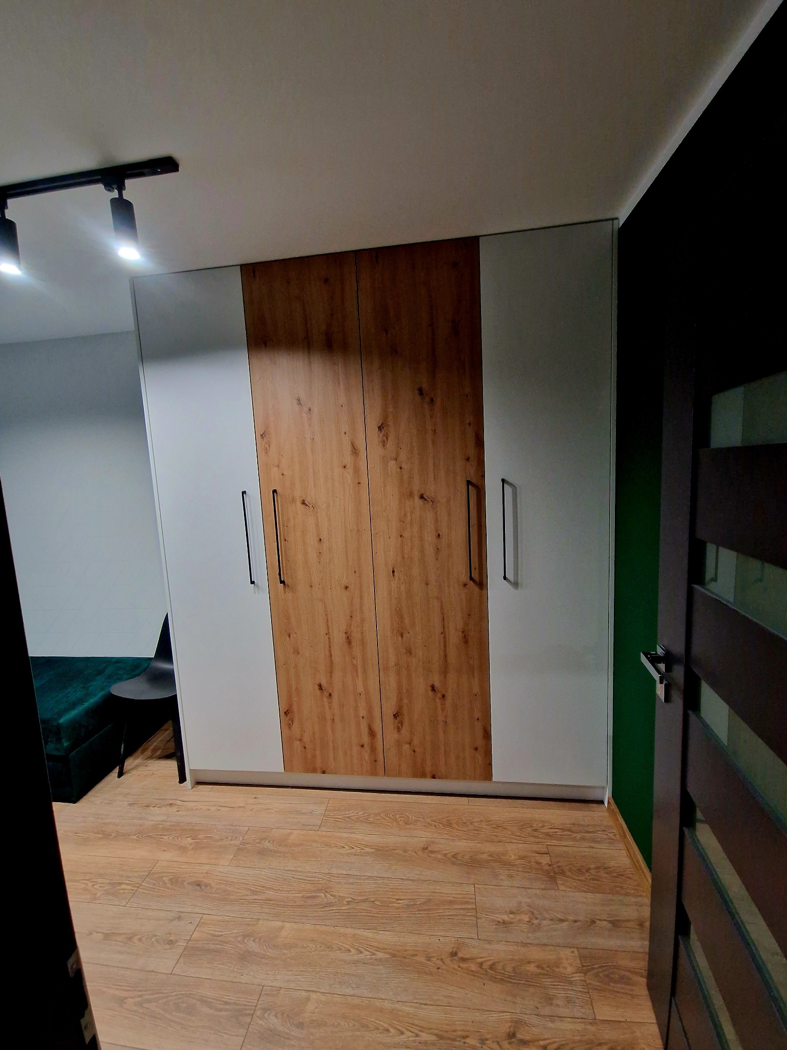 Nowoczesna szafa z frontami łączącymi biel i dekor drewna, z długimi, czarnymi uchwytami, na tle drewnianej podłogi i fragmentu zielonej ściany.