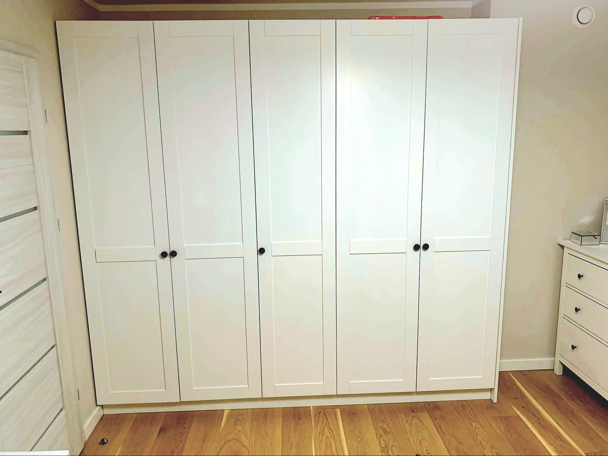 Biała szafa z sześcioma drzwiami z czarnymi uchwytami, na drewnianej podłodze. Uwagę przyciąga minimalistyczny design i dopasowanie do wnętrza.