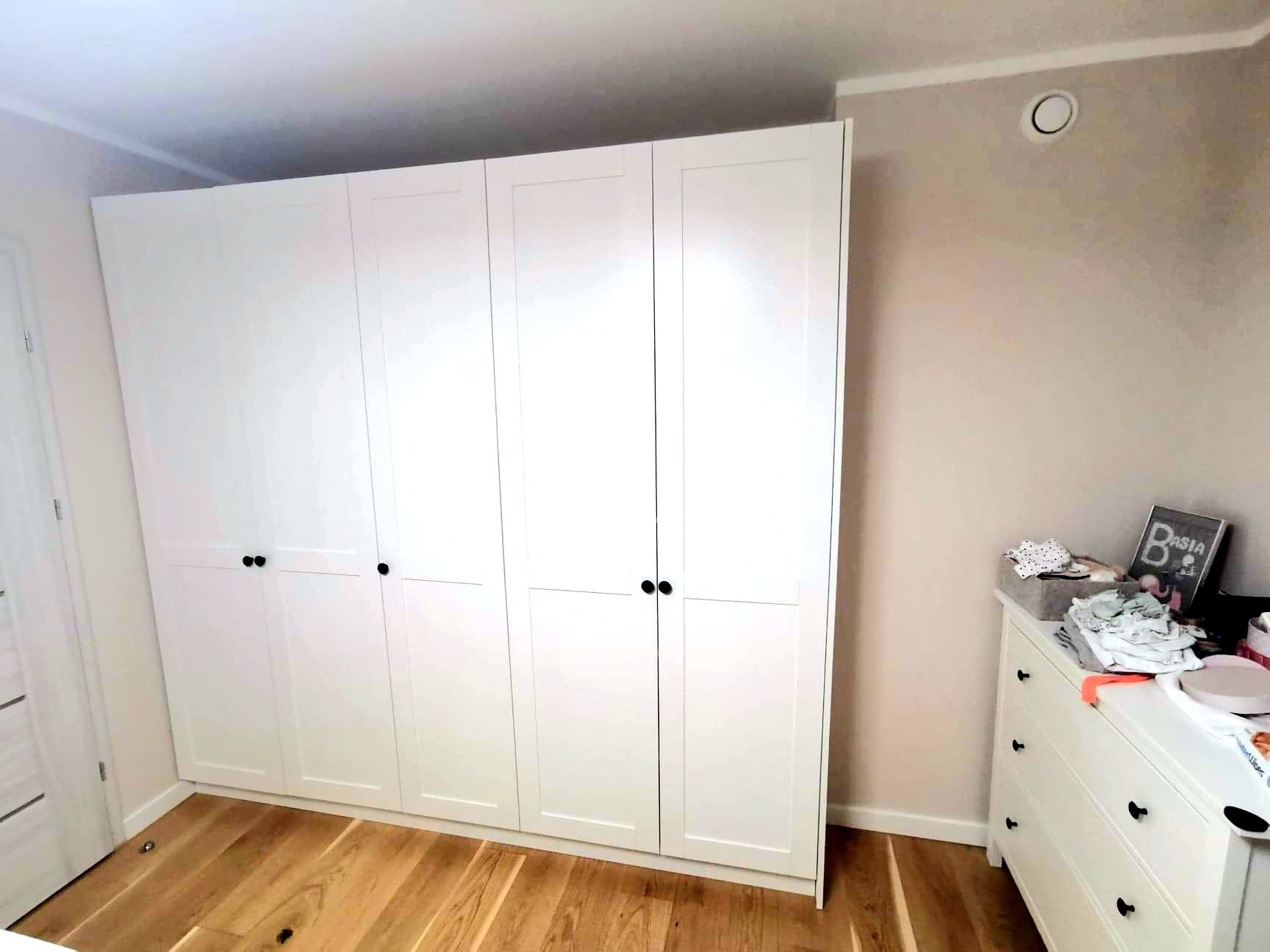 Biała szafa z czterema drzwiami i czarnymi uchwytami, na drewnianej podłodze, w pokoju z komodą i dekoracjami. Minimalistyczny design, jasne wnętrze.