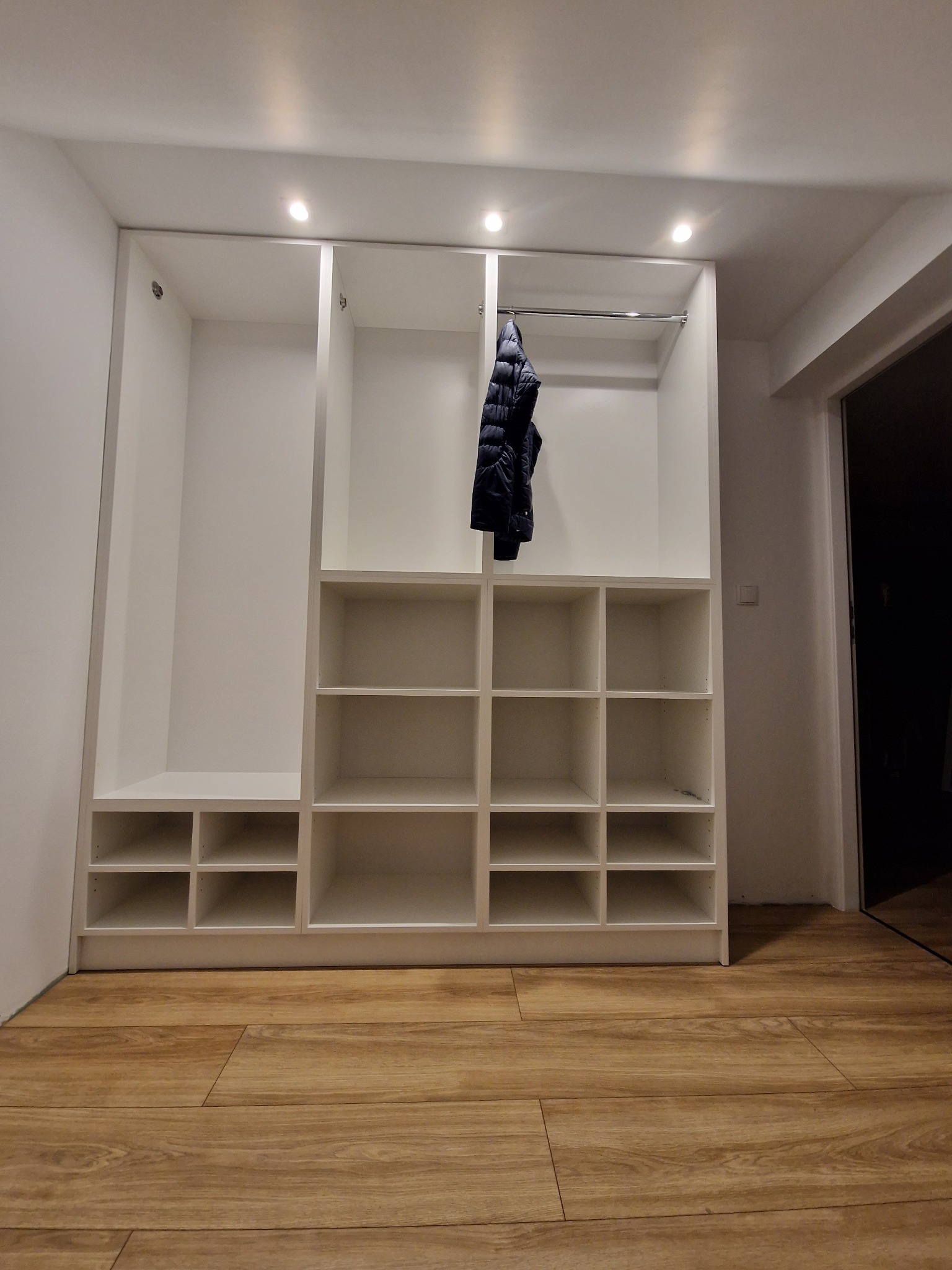 Biała, otwarta szafa wnękowa z półkami i drążkiem, oświetlona punktowo, z kurtką wiszącą na drążku, na podłodze panele imitujące drewno, widok z dołu.