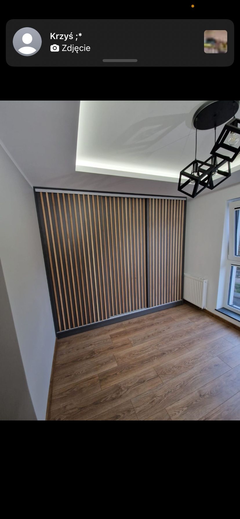 Nowoczesna szafa wnękowa z przesuwnymi drzwiami lamelowymi w jasnym wnętrzu. Widoczne drewniane panele i minimalistyczna lampa.