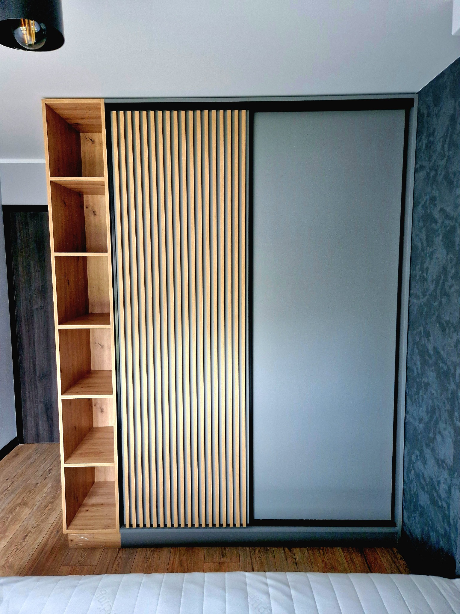 Nowoczesna szafa wnękowa z przesuwnymi drzwiami: matowe szkło i drewniane lamele. Obok regał z jasnego drewna. Minimalistyczny design, szara ściana w tle.
