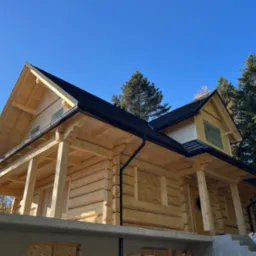 Surowy stan drewnianego domu z bali z czarnym dachem, widoczne deski zakrywające okna i szara podmurówka, w tle drzewa iglaste i błękitne niebo.