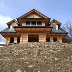 Surowy stan budowy domu z bali, z widoczną konstrukcją drewnianą, czarnym dachem i przygotowanym terenem pod budowę na tle błękitnego nieba.